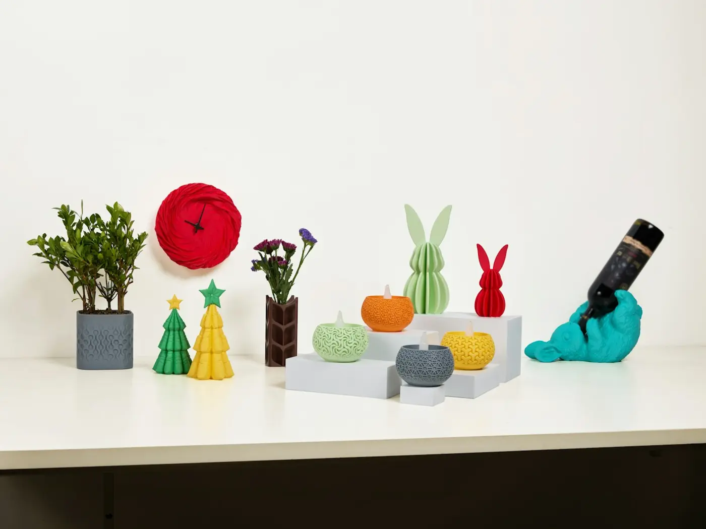Peças decorativas impressas em 3D: relógio, árvores de Natal, velas e coelhos coloridos sobre uma bancada.